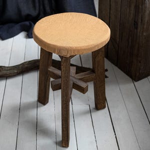 Puede incluir: Un pequeño taburete redondo de madera con asiento marrón claro y patas marrones oscuras. Las patas están conectadas por un soporte en forma de cruz. El taburete está sobre un suelo de madera pintado de blanco.