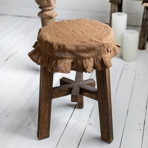Pu&ograve; includere: Uno sgabello in legno marrone con un sedile rotondo rivestito in tessuto marrone chiaro con volant. Lo sgabello ha quattro gambe robuste e una struttura di supporto a forma di croce. Lo sgabello &egrave; posto su un pavimento di legno bianco.