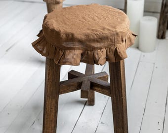 May include: A brown wooden stool with a round seat covered in a ruffled, light brown fabric. The stool has four sturdy legs and a cross-shaped support structure. The stool is placed on a white wooden floor.