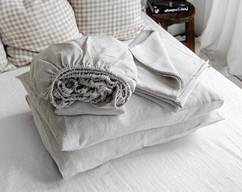 Puede incluir: Una cama con una sábana blanca y varias almohadas. Las almohadas incluyen patrones grises claros lisos y a cuadros. Una sábana bajera y un juego de sábanas dobladas con finas rayas grises están apiladas sobre las almohadas.