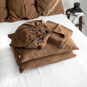 May include: A set of brown linen bedding, including a fitted sheet, flat sheet, and two pillowcases. The bedding is arranged on a white bedspread.