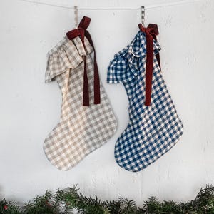 Könnte beinhalten: Zwei Weihnachtsstrümpfe hängen an einer weißen Wand. Einer ist beige und weiß kariert, der andere blau und weiß kariert. Beide haben Rüschen und bordeauxfarbene Samtbänder. Grüne Girlande mit roten Beeren schmückt den unteren Bereich.
