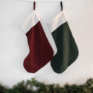 Pode incluir: Duas meias de Natal penduradas contra uma parede branca. Uma é bordô, a outra verde escuro, ambas com punhos brancos. Uma grinalda de galhos de pinheiro verde com bagas vermelhas decora a parte inferior.