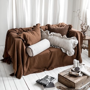 Peut inclure: Un canapé marron recouvert d'un plaid assorti et orné de plusieurs coussins. Les coussins comprennent un coussin à volants, un coussin marron texturé et un traversin blanc avec une bordure noire. La scène se déroule devant un rideau blanc.