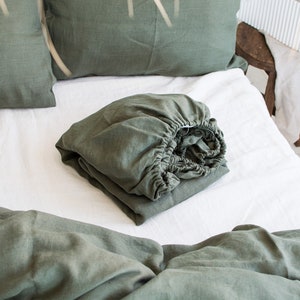 Op de afbeelding: Een gevouwen saliegroen linnen hoeslaken op een wit bed met een saliegroen linnen kussen erop.