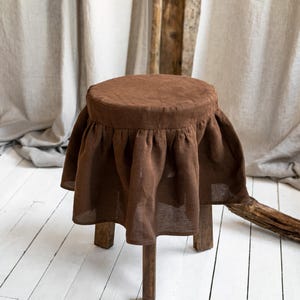 May include: A small, wooden stool with a round, brown upholstered seat. The seat has a gathered fabric skirt, giving it a vintage or farmhouse aesthetic. The stool's legs are simple and sturdy, made of natural wood. The stool is a charming piece.