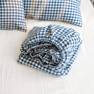 May include: A folded blue and white gingham fitted sheet and two matching pillowcases on a white sheet. The bedding set is made of a soft fabric, ideal for a bedroom.