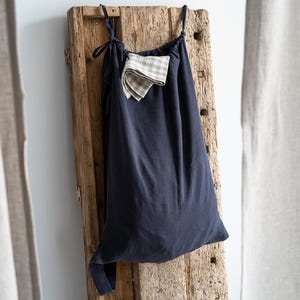 Può includere: Una borsa blu navy con coulisse con un piccolo panno a quadretti piegato che spunta. La borsa è appesa a una tavola di legno stagionato. Il tessuto della borsa appare morbido e leggermente stropicciato. Il cordoncino è legato in alto.