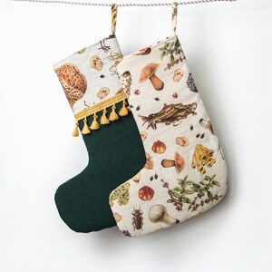May include: A green and cream Christmas stocking with a mushroom and forest-themed print. The stocking has a gold tassel and is hanging from a string.