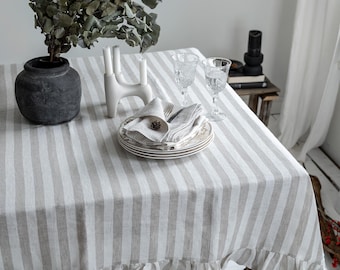 May include: A linen tablecloth with vertical gray and white stripes and a ruffled edge. The table is set with plates, napkins, and glassware. A decorative vase with greenery and a white candle holder are also on the table.