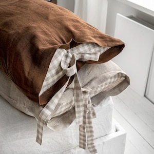 May include: Close-up of two decorative pillows. The top pillow is a solid brown color, and the bottom pillow is a light beige. The pillowcases have a gingham pattern on the inside and are tied with matching fabric ties. The pillows are on a white bed.