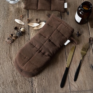 May include: A brown quilted oven mitt and matching square pot holders are displayed on a wooden surface, alongside a knife, fork, and a candle in a glass jar. The mitt and pot holders have a stitched grid pattern.