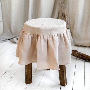 Puede incluir: Un pequeño taburete redondo de madera con una funda de lino rosa claro y una falda con volantes. El taburete tiene cuatro patas de color marrón oscuro y se apoya sobre un suelo de madera pintado de blanco. El fondo presenta cortinas de color neutro.