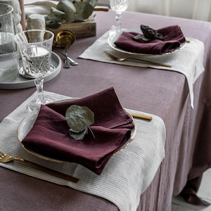 Peut inclure: Une table de salle à manger dressée pour un repas. La table est recouverte d'une nappe mauve et garnie d'assiettes, de serviettes et de verres. Les serviettes sont de couleur bordeaux foncé et les assiettes ont des bords dorés. L'ensemble comprend des fourchettes dorées.