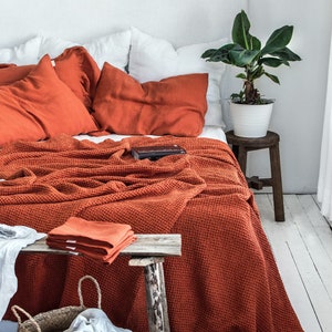 Könnte beinhalten: Eine orangefarbene Waffel-Decke liegt auf einem weißen Bett mit orangefarbenen Leinenkissen. Ein kleiner Holzhocker mit einem weißen Topf und einer grünen Pflanze steht neben dem Bett.