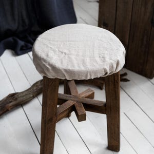 Puede incluir: Un pequeño taburete de madera rústico con una cubierta de asiento redonda de lino claro. El taburete tiene cuatro patas robustas de color marrón oscuro y una estructura de soporte en forma de cruz. El fondo presenta una tabla de madera y una tela azul oscuro.