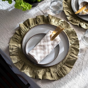 Può includere: Un'apparecchiatura con un sottopiatto verde chiaro a balze, un piatto grigio e un tovagliolo a quadretti piegato. Forchetta e coltello color oro sono posizionati sul tovagliolo. Sono visibili anche una ciotola di prodotti freschi e un bicchiere d'acqua.