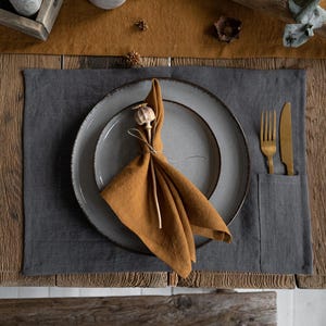 Pode incluir: Vista aérea de uma mesa posta com um tapete cinza, pratos cinza e uma toalha de mesa mostarda dobrada. Garfo e faca dourados em um bolso no tapete. Uma vela e itens decorativos no fundo.