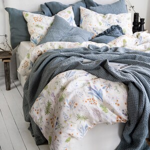 May include: A bed with a white headboard and bedding. The bed has several blue pillows and a blue waffle-weave blanket. The duvet cover and pillowcases feature a floral pattern with green, blue, and orange accents. The room has white painted wood floors.