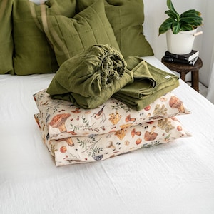 May include: A stack of green linen pillowcases and a fitted sheet with a mushroom and bird print. The bedding is on a white bed with green pillows.