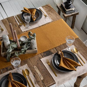Op de afbeelding: Een rustieke houten tafel gedekt voor een maaltijd. De tafel is versierd met mosterdkleurige servetten, placemats en een tafelloper. Elke plaatsing omvat een donker bord, gouden bestek en een waterglas. Een houten dienblad bevat kaarsen en groen.