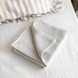 May include: Folded, striped bedding set in neutral tones. The set includes a folded blanket or sheet with thin, light gray stripes, and a pillowcase with wider stripes and a ruffled edge. The bedding is made of a soft, natural fabric.