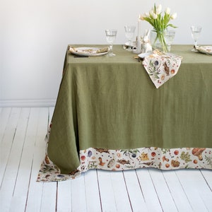 Puede incluir: Un mantel de lino verde con un borde de estampado floral blanco y marrón. El mantel está puesto con dos cubiertos, cada uno con un plato blanco, un vaso y una servilleta. Una figurita de conejo de cerámica blanca se encuentra en la mesa, y un jarrón de flores blancas está en el centro.