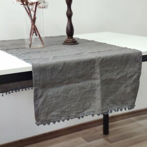 May include: A gray linen table runner with a pom-pom trim. The runner is on a white table with a wooden candlestick and a vase of cotton flowers.