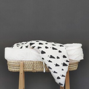 May include: A white wicker bassinet with a white and black patterned blanket inside. The bassinet is on a wooden stand with two legs.