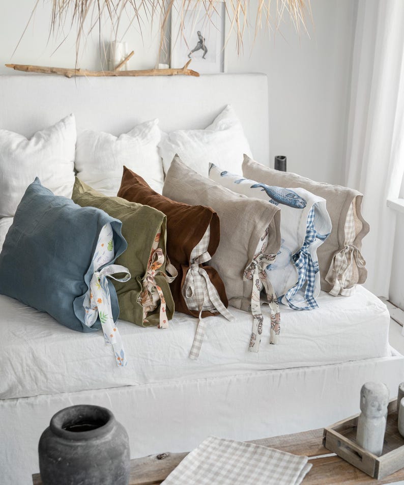 May include: A collection of linen pillowcases in various colors, including blue, green, and brown, with patterned ties. Some pillowcases feature printed designs, such as a whale. The pillows are arranged on a white bed, creating a cozy and inviting atmosphere.