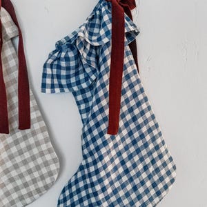 May include: Two Christmas stockings hanging against a white wall. One stocking is blue and white gingham, the other is gray and white gingham. Both have a burgundy ribbon and bow.