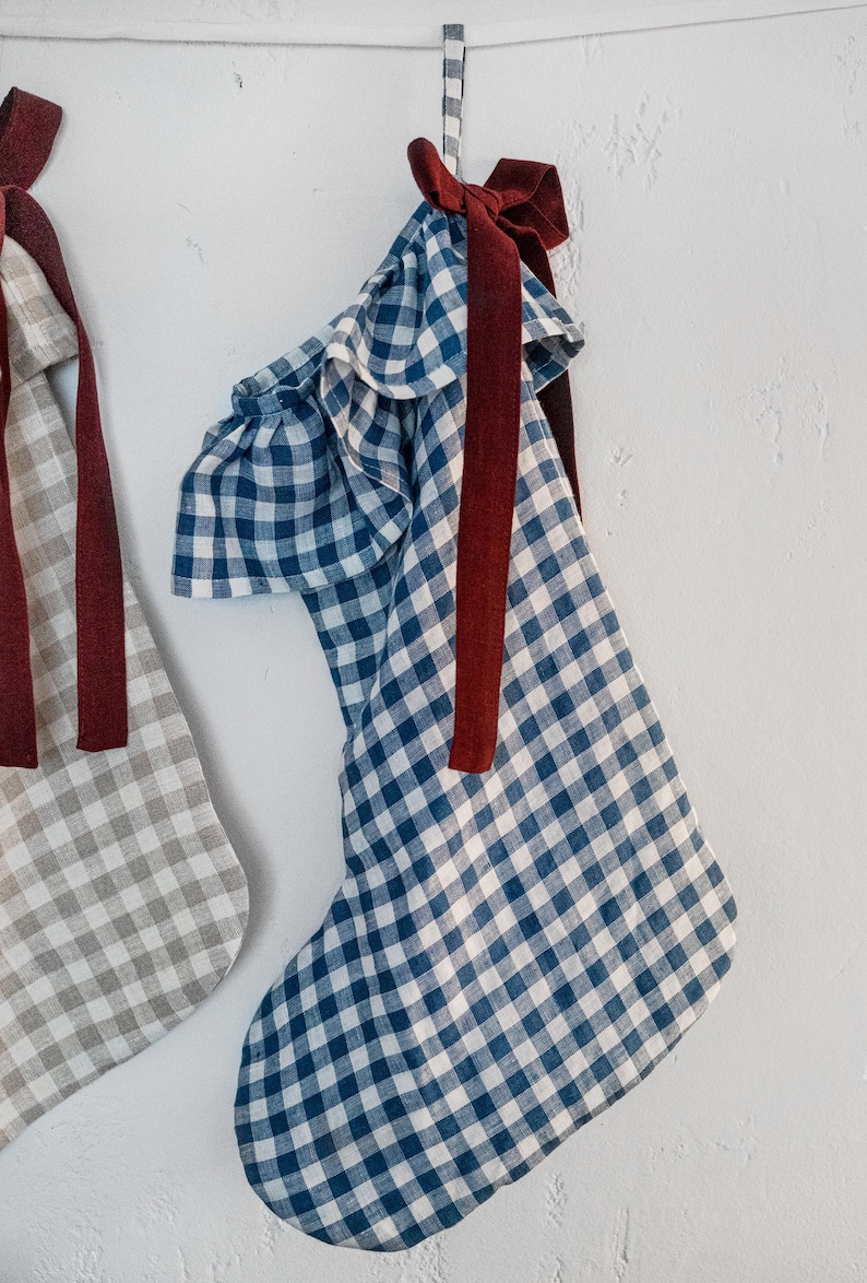 May include: Two Christmas stockings hanging against a white wall. One stocking is blue and white gingham, the other is gray and white gingham. Both have a burgundy ribbon and bow.
