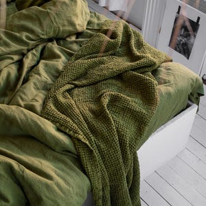 Puede incluir: Una manta de tejido de gofre verde drapeada sobre una cama con sábanas de lino verde a juego.