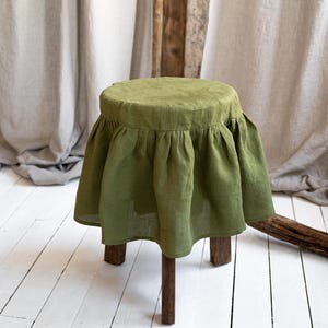May include: A small, wooden stool with a round, olive green linen cover. The cover has a ruffled skirt detail. The stool has three dark brown legs and sits on a white-painted wooden floor. The background includes neutral-colored curtains and wooden beams.