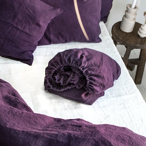 May include: A purple linen fitted sheet is folded and placed on a white linen bedspread. Two purple linen pillowcases are on the bed.