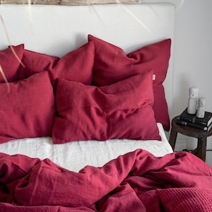 May include: A red linen duvet cover and pillowcases are arranged on a bed with a white headboard. The bedding is rumpled and casual, creating a cosy and inviting atmosphere.