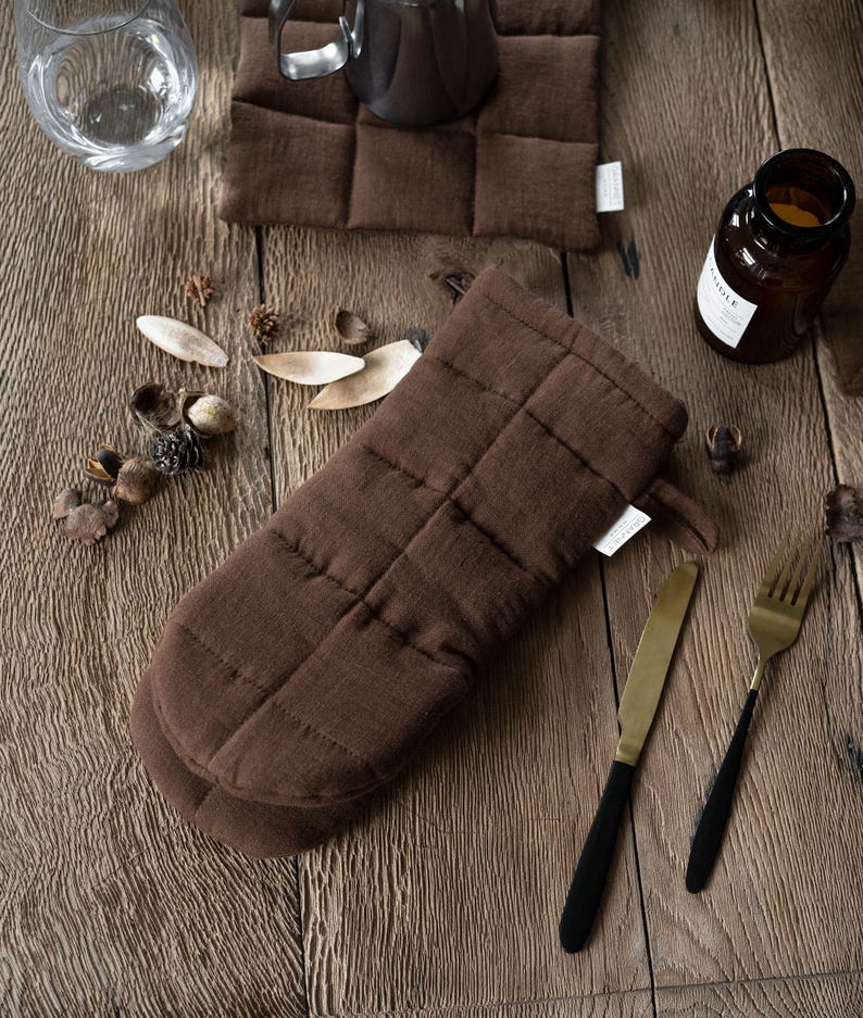 May include: A brown quilted oven mitt and matching square pot holders are displayed on a wooden surface, alongside a knife, fork, and a candle in a glass jar. The mitt and pot holders have a stitched grid pattern.
