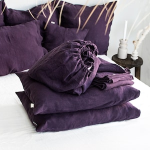 Puede incluir: Una pila de fundas de almohada de lino morado y una funda de almohada. Las fundas de almohada están dobladas y apiladas una encima de la otra. La funda de almohada está en la parte superior de la pila de fundas de almohada. Las fundas de almohada y la funda de almohada están hechas de una tela de lino suave y natural.