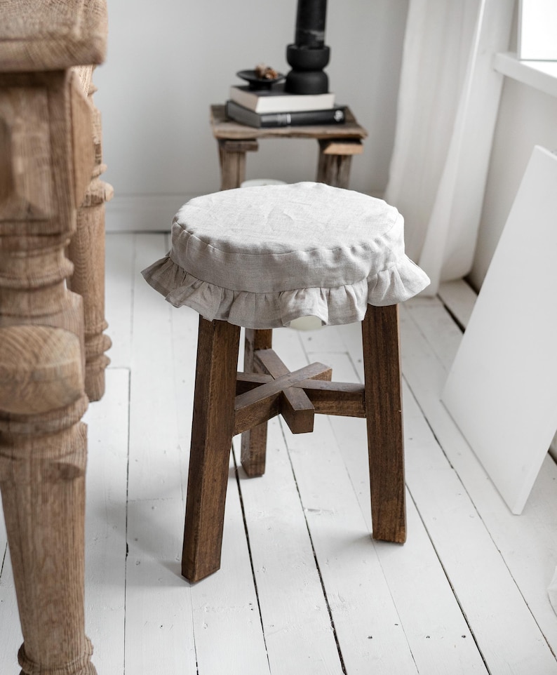 Pu&ograve; includere: Uno sgabello in legno con una seduta rotonda in lino chiaro con un bordo a balze. Lo sgabello ha una struttura in legno rustico marrone scuro con un supporto a forma di croce. Lo sfondo comprende un tavolino in legno e assi del pavimento verniciate di bianco.