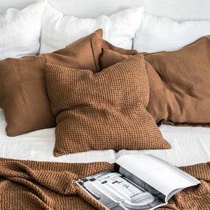 May include: A brown waffle weave throw blanket is folded on a bed with white linen bedding. The blanket is textured and has a waffle pattern. There are brown linen pillows on the bed, and a magazine is lying on top of the blanket.