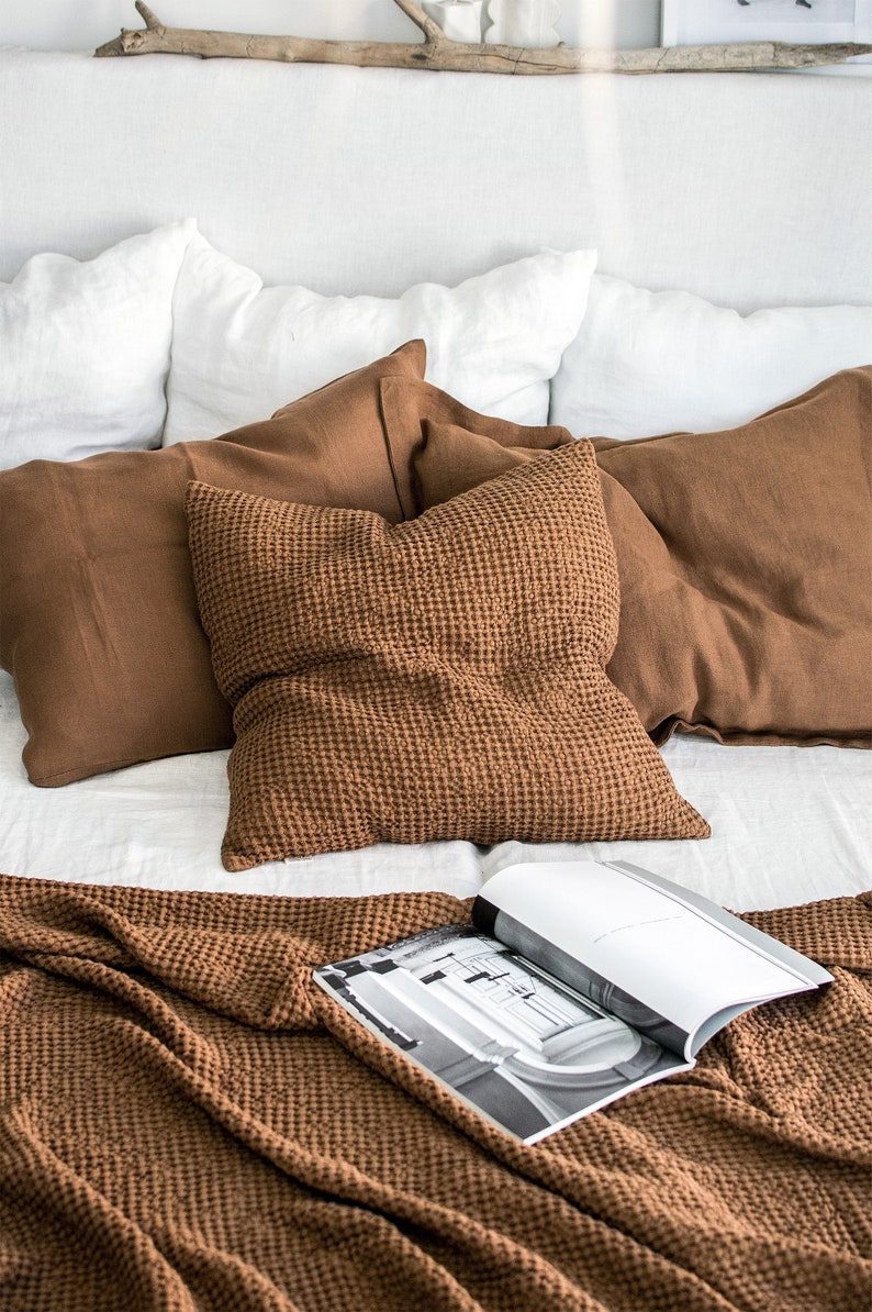 May include: A brown waffle weave throw blanket is folded on a bed with white linen bedding. The blanket is textured and has a waffle pattern. There are brown linen pillows on the bed, and a magazine is lying on top of the blanket.