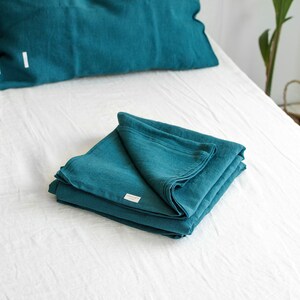 Könnte beinhalten: Ein gefalteter Stapel tealfarbener Leinenstoff auf einem weißen Bett. Der Stoff ist ordentlich gefaltet und hat ein kleines weißes Etikett mit dem Wort "LINEN" darauf.