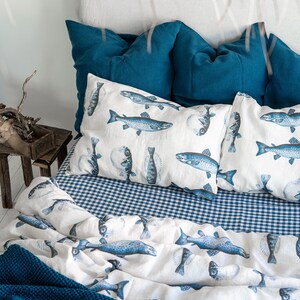 May include: A bed with a white headboard and teal pillows. The bedding features a white duvet and pillowcases printed with blue fish illustrations. A blue and white gingham sheet and a teal blanket complete the nautical theme.
