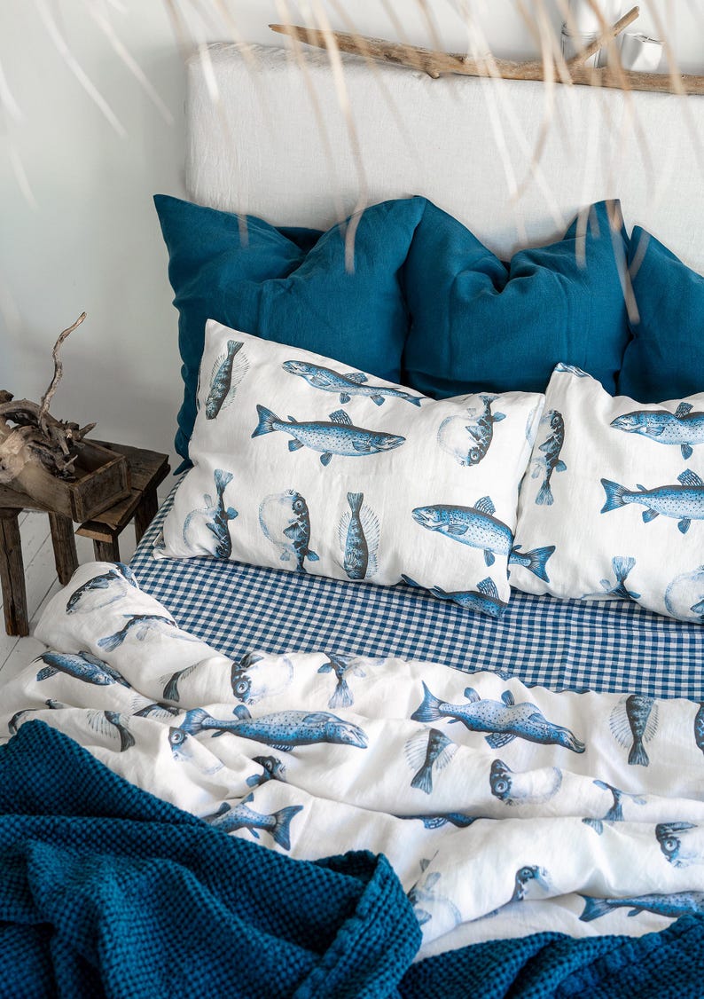 May include: A bed with a white headboard and teal pillows. The bedding features a white duvet and pillowcases printed with blue fish illustrations. A blue and white gingham sheet and a teal blanket complete the nautical theme.