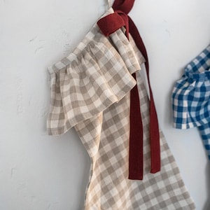 May include: A beige and white gingham Christmas stocking with a ruffled top and a burgundy ribbon. The stocking is hanging against a white wall. Another blue and white gingham stocking is partially visible.