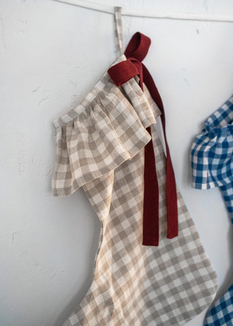 May include: A beige and white gingham Christmas stocking with a ruffled top and a burgundy ribbon. The stocking is hanging against a white wall. Another blue and white gingham stocking is partially visible.