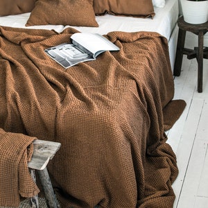 May include: A brown waffle weave throw blanket is draped over a bed with brown linen pillows. The blanket is folded at the foot of the bed and hangs over the side. A magazine is placed on top of the blanket.