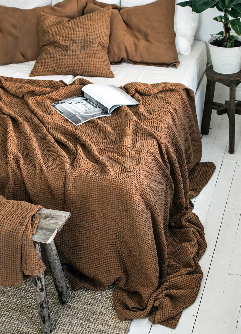May include: A brown waffle weave throw blanket is draped over a bed with brown linen pillows. The blanket is folded at the foot of the bed and hangs over the side. A magazine is placed on top of the blanket.
