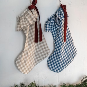 May include: Two Christmas stockings hang against a white wall. One is tan and white gingham, the other is blue and white gingham. Both have a ruffled top and a burgundy ribbon. Greenery with red berries is at the bottom.