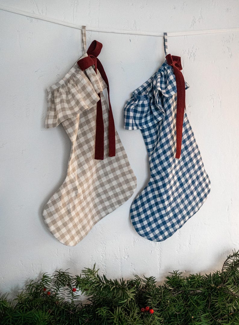 May include: Two Christmas stockings hang against a white wall. One is tan and white gingham, the other is blue and white gingham. Both have a ruffled top and a burgundy ribbon. Greenery with red berries is at the bottom.